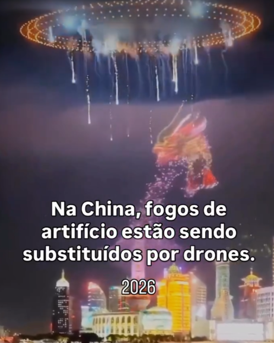 Imagem mostra uma cena noturna com um espetáculo aéreo sobre uma cidade à beira de um rio ou mar, com prédios iluminados ao fundo e reflexos das luzes na água. No céu, há formações circulares luminosas e efeitos verticais semelhantes a cascatas de luz, além da figura de um dragão formada por pontos luminosos em tons de vermelho, laranja e amarelo. Sobre a imagem, aparece um texto em letras brancas que diz ‘Na China, fogos de artifício estão sendo substituídos por drones.’ e, logo abaixo, o número ‘2026’, sem a presença de pessoas visíveis em primeiro plano.
