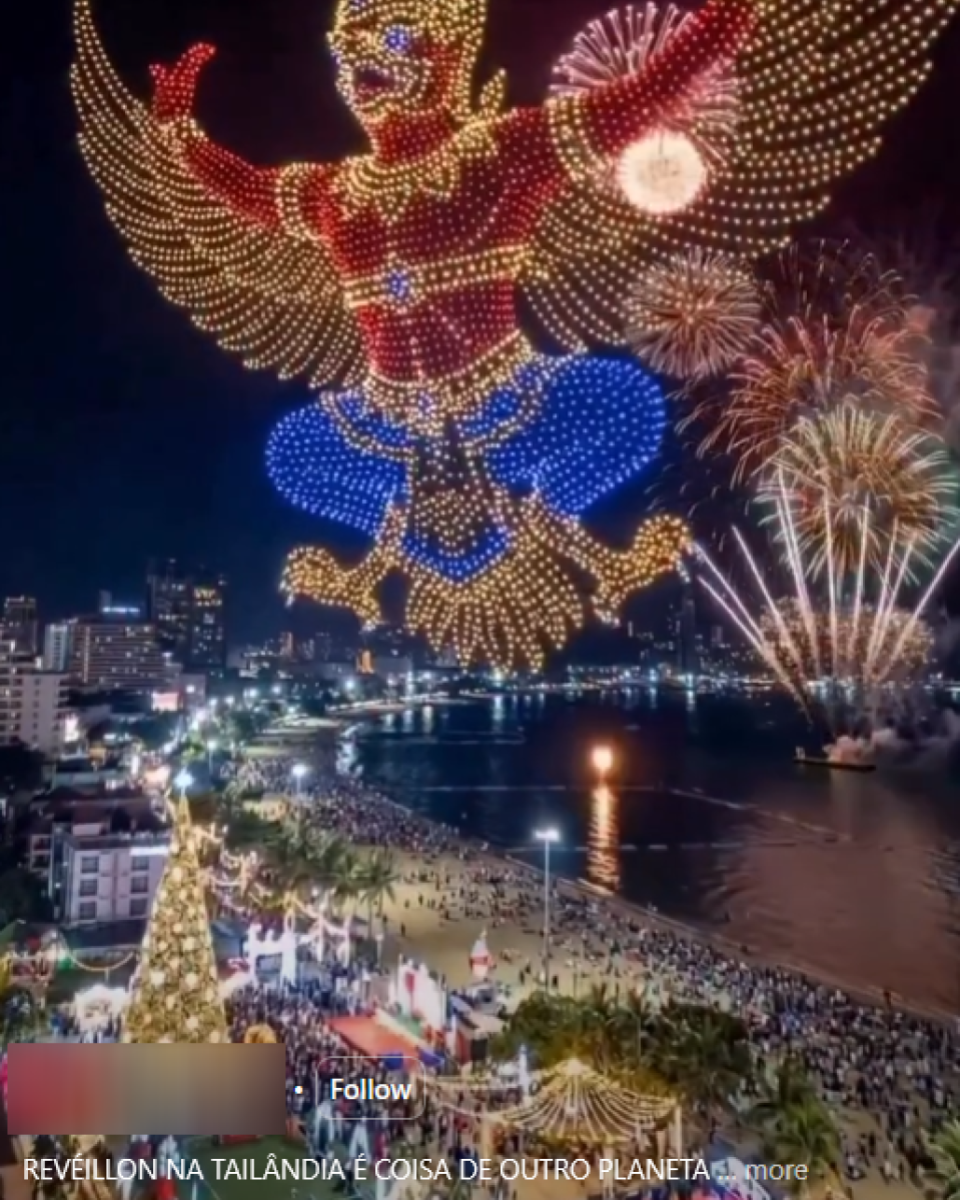 Imagem mostra uma cena noturna à beira-mar com um grande show de drones formando no céu a figura de uma entidade alada humanoide, composta por pontos de luz nas cores dourado, vermelho e azul, com asas abertas e braços erguidos. Ao fundo, há fogos de artifício explodindo sobre a água, refletindo no mar, e uma orla iluminada com prédios, árvores e uma multidão concentrada ao longo da praia e do calçadão. Na parte inferior, é possível ver estruturas decorativas iluminadas e um texto sobreposto que menciona ‘REVÉILLON NA TAILÂNDIA’, além de elementos gráficos típicos de uma publicação em rede social.