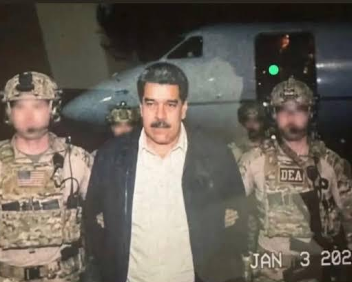 A imagem mostra Nicolás Maduro ao centro, de cabelo escuro e bigode, vestindo uma camisa clara por baixo de um casaco escuro, caminhando ou sendo conduzido. Ele está ao lado de dois homens usando uniformes militares camuflados, coletes táticos e capacetes. O homem à esquerda tem o rosto desfocado e porta equipamentos presos ao colete. O homem à direita também está com o rosto desfocado e traz a inscrição ‘DEA’ visível em seu colete. Ambos aparentam estar segurando ou acompanhando o homem ao centro. Ao fundo, há outros agentes parcialmente visíveis, também com trajes militares, e a lateral de uma aeronave de cor clara, com uma porta aberta e uma luz verde acesa próxima à entrada. No canto inferior direito da imagem aparece uma marcação de data com os dizeres ‘JAN 3 202…’, com os últimos dígitos do ano cortados ou fora do enquadramento. A cena aparenta ocorrer em ambiente noturno ou com pouca iluminação.
