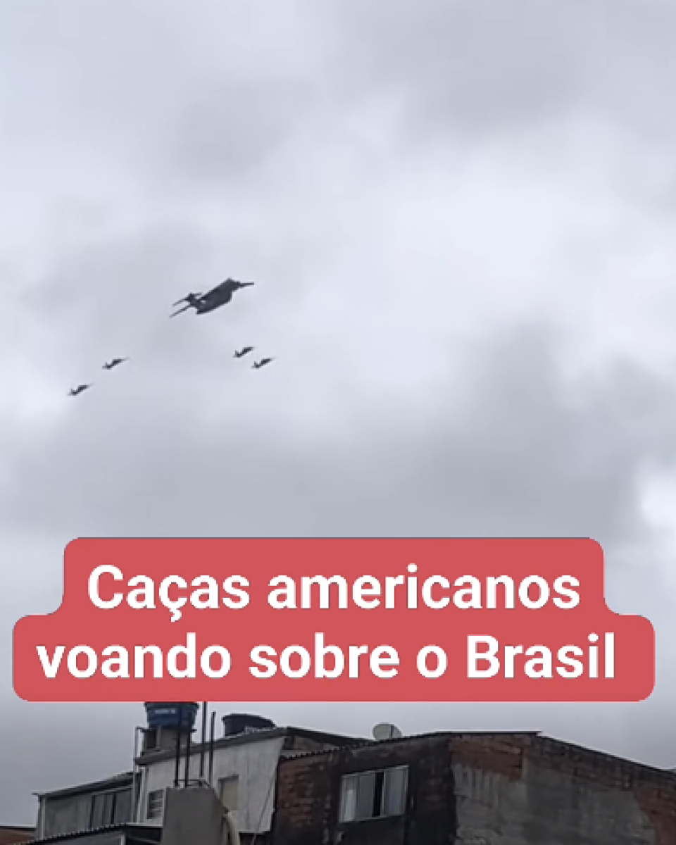 A imagem mostra um céu nublado, com nuvens claras e acinzentadas ocupando quase todo o enquadramento. Na parte superior esquerda, aparecem cinco aeronaves voando em formação: uma aeronave maior à frente e outras menores distribuídas ao redor, todas vistas à distância. Na parte inferior da imagem, vê-se o topo de construções urbanas, com telhados, paredes de alvenaria, uma caixa-d’água azul e algumas antenas e equipamentos instalados sobre os prédios. Sobre a imagem, ao centro, há um retângulo arredondado vermelho com texto branco escrito: ‘Caças americanos voando sobre o Brasil’.