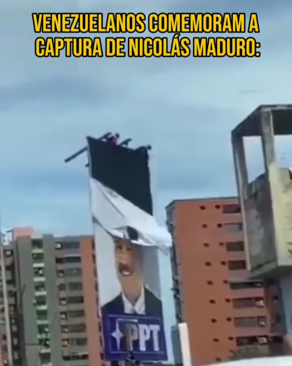 Imagem mostra ambiente urbano durante o dia, com prédios residenciais de vários andares ao fundo, em tons de bege, cinza e laranja. No centro da cena, há um outdoor vertical fixado sobre uma estrutura elevada, exibindo parcialmente a imagem de Nicolás Maduro — homem de pele morena, cabelo escuro e bigode, vestindo terno escuro e camisa clara, com um logotipo e a sigla ‘PPT’ na parte inferior; o painel aparece rasgado ou sendo retirado na parte superior, com uma lona branca solta e dobrada. No topo da imagem, há um texto sobreposto em letras grandes e amarelas que diz ‘VENEZUELANOS COMEMORAM A CAPTURA DE NICOLÁS MADURO’.