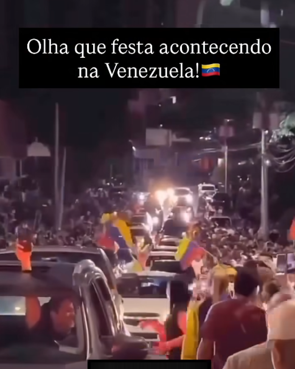 Imagem mostra uma cena noturna em uma via urbana tomada por uma multidão, com muitas pessoas de diferentes idades e gêneros, vestindo camisetas de mangas curtas e roupas casuais, reunidas entre carros parados ou em movimento lento. Algumas pessoas aparecem com parte do corpo para fora dos veículos, segurando bandeiras da Venezuela nas cores amarelo, azul e vermelho, enquanto outras caminham ao lado dos automóveis. A rua é iluminada por postes de luz e faróis, com prédios baixos e fiação elétrica visíveis ao fundo, e há um texto sobreposto em fundo preto na parte superior da imagem que diz ‘Olha que festa acontecendo na Venezuela!’ acompanhado de um emoji da bandeira venezuelana.