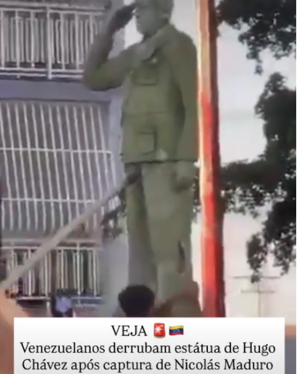 Imagem mostra uma cena diurna em área urbana, com estátua de um homem em posição ereta, vestindo uniforme militar de cor verde-oliva, com boina e uma das mãos levantadas em gesto de continência. A escultura está instalada sobre um pedestal, próxima a uma estrutura com grades metálicas e árvores ao fundo, enquanto pessoas aparecem parcialmente visíveis na base da estátua. Na parte inferior da imagem, há uma tarja branca com o logotipo ‘VEJA’, acompanhada de ícones gráficos, e um texto que diz ‘Venezuelanos derrubam estátua de Hugo Chávez após captura de Nicolás Maduro’.