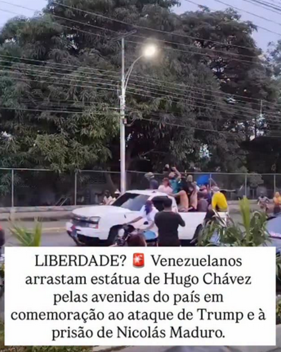 Imagem mostra uma cena diurna em uma avenida urbana arborizada, com uma caminhonete branca em movimento lento no centro da via, cercada por várias pessoas. Sobre a carroceria do veículo, há homens e mulheres de diferentes idades, majoritariamente de pele morena e clara, vestindo roupas casuais como camisetas e shorts, alguns sentados e outros em pé, com braços erguidos. Ao redor da caminhonete, outras pessoas caminham ou observam, enquanto postes de iluminação, fiação elétrica, uma cerca metálica e árvores de grande porte aparecem ao fundo. Na parte inferior da imagem, há uma tarja branca com texto em letras pretas que diz ‘LIBERDADE? Venezuelanos arrastam estátua de Hugo Chávez pelas avenidas do país em comemoração ao ataque de Trump e à prisão de Nicolás Maduro’.