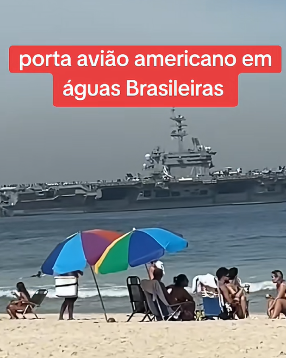 Imagem mostra cena diurna em praia, com areia clara em primeiro plano e várias pessoas sentadas ou em pé próximas à beira-mar, usando roupas de banho, cadeiras de praia e guarda-sóis. Ao fundo, no mar, aparece uma grande embarcação militar de cor cinza, com estrutura elevada, antenas e pista de pouso visíveis, navegando paralelamente à costa. Na parte superior da imagem, há um texto sobreposto em fundo vermelho que diz: ‘porta avião americano em águas Brasileiras’.