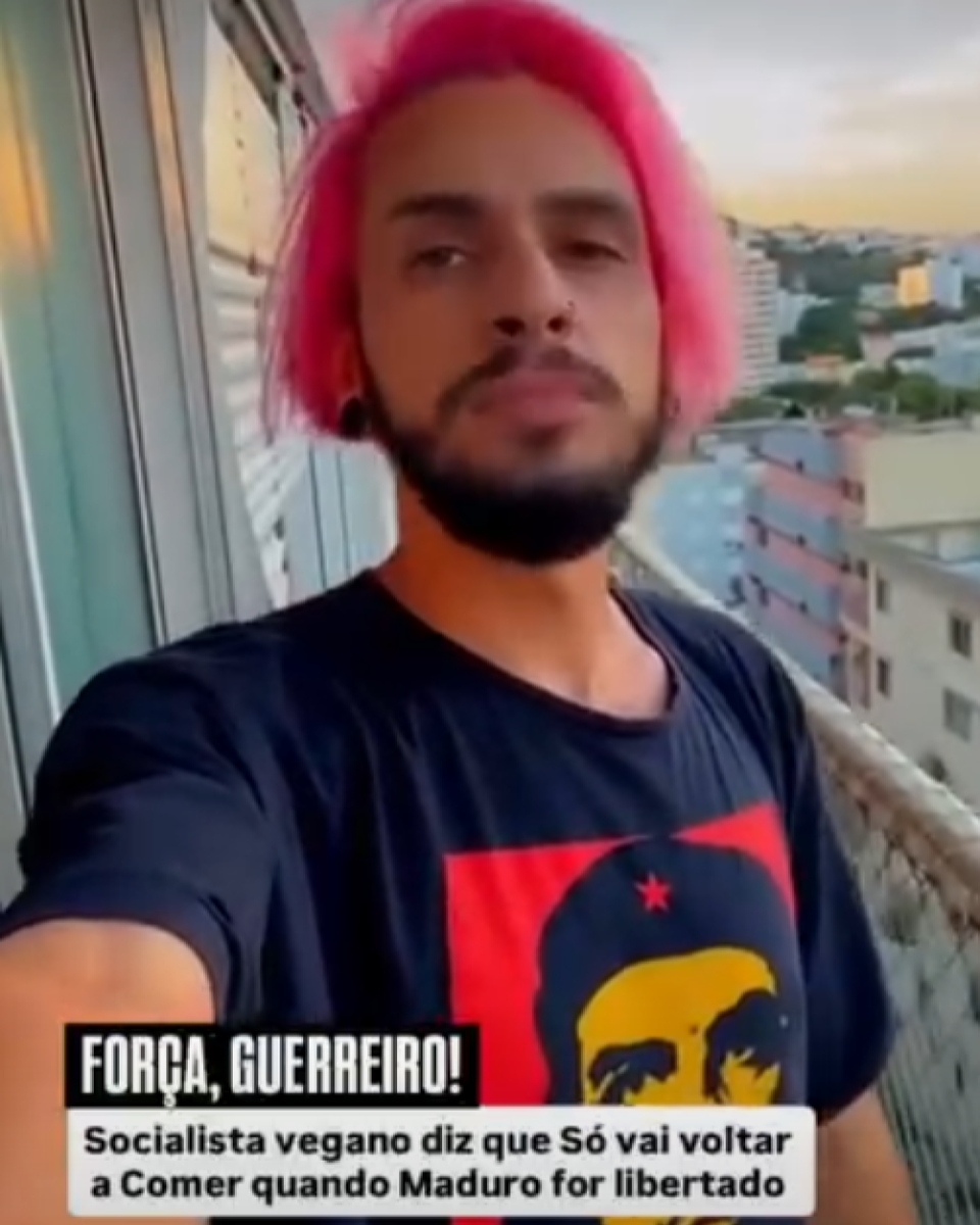 Imagem mostra homem jovem de pele morena, cabelo médio tingido de rosa e barba escura curta, filmando a si mesmo em uma varanda, com prédios residenciais e o céu ao entardecer ao fundo. Ele veste camiseta preta com uma estampa em vermelho, amarelo e preto que traz o rosto estilizado de Che Guevara e uma estrela vermelha. Na parte inferior da imagem, há uma tarja com texto que diz ‘FORÇA, GUERREIRO!’ e, abaixo, ‘Socialista vegano diz que só vai voltar a comer quando Maduro for libertado’.