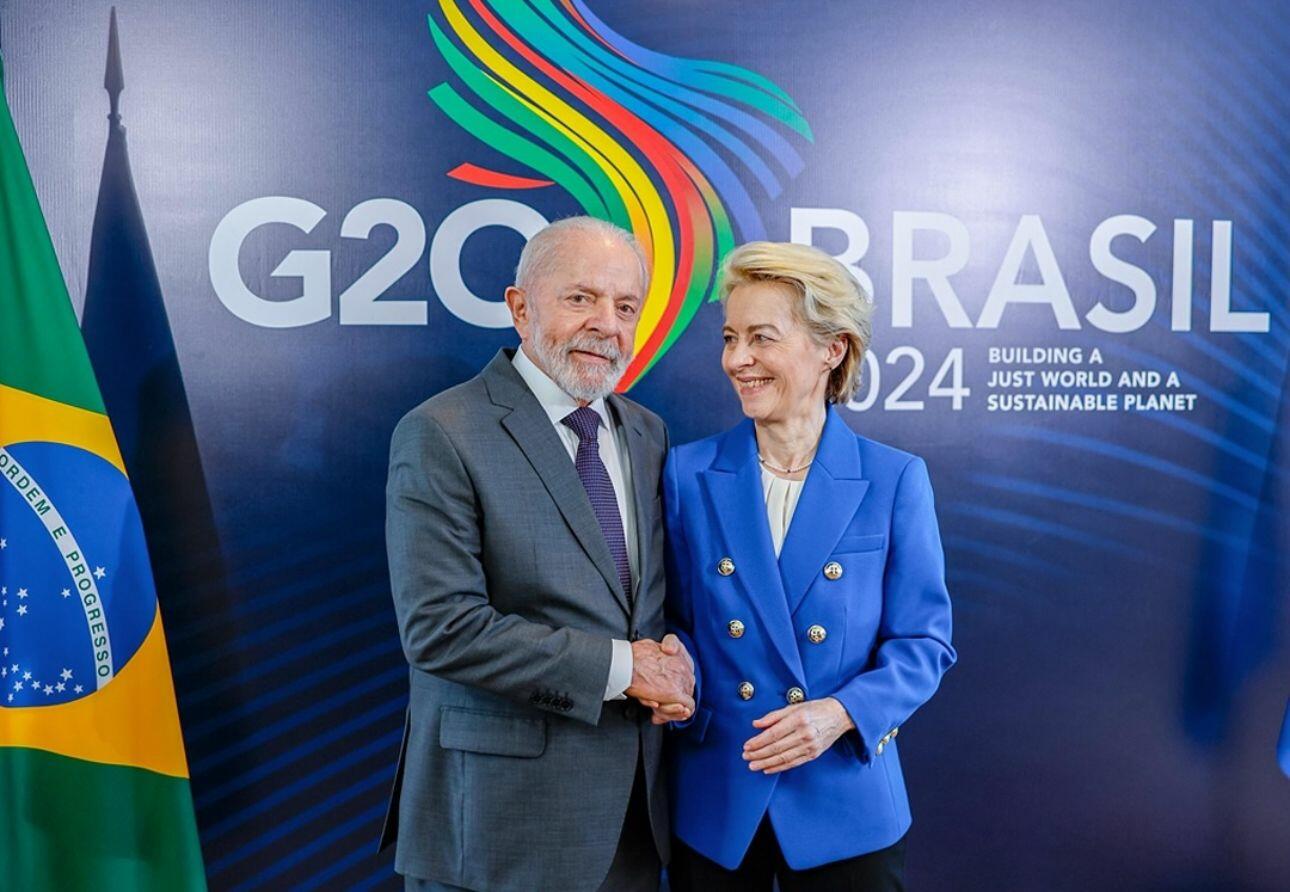 Imagem de duas pessoas em pé, posicionadas lado a lado, diante de um painel institucional. À esquerda, há uma bandeira do Brasil parcialmente visível, com as cores verde, amarelo, azul e branco. No centro da imagem está Lula, um homem com cabelo curto grisalho e barba branca, vestindo um terno cinza, camisa branca e gravata escura. Ele segura a mão da pessoa ao seu lado. À direita está Ursula von der Leyen, uma mulher com cabelo curto loiro, vestindo um conjunto azul composto por blazer com botões dourados e blusa clara por baixo. Ambos estão voltados para a frente, com os corpos levemente inclinados um em direção ao outro. Ao fundo, o painel exibe a inscrição ‘G20 BRASIL 2024’ e, abaixo, o texto ‘Building a Just World and a Sustainable Planet’, além de um elemento gráfico colorido em forma de linhas curvas nas cores verde, amarelo, vermelho e azul. O fundo é predominantemente azul escuro, com iluminação uniforme.