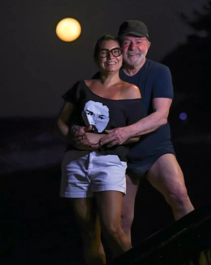 Imagem mostra o presidente Lula e a primeira-dama Janja em um ambiente externo noturno, com a lua cheia visível ao fundo. Lula é um homem de pele clara, cabelos e barba curtos e grisalhos, que usa uma camiseta azul-escura, boné da mesma cor e trajes de banho escuros, aparecendo em pé atrás de Janja com as mãos sob o abdômen dela. Janja é uma mulher de pele clara, cabelos escuros presos, que usa óculos de grau com armação escura, uma camiseta preta com uma estampa de rosto humano em tons de cinza e shorts brancos. Ambos estão sorrindo para a câmera em um plano médio, com a iluminação focada neles e o restante do cenário em sombras.