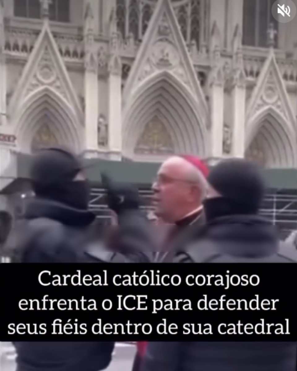 Imagem mostra vídeo em ambiente externo, diante da fachada clara de uma catedral com arcos altos e detalhes góticos. No centro, aparece um homem idoso de pele clara, calvo, usando solidéu vermelho e veste religiosa escura, visto de perfil, cercado por pessoas com roupas pretas e rostos desfocados nas laterais, sugerindo agentes ou seguranças. Na parte inferior, sobre uma faixa preta, há um texto em letras brancas que diz: ‘Cardeal católico corajoso enfrenta o ICE para defender seus fiéis dentro de sua catedral’.