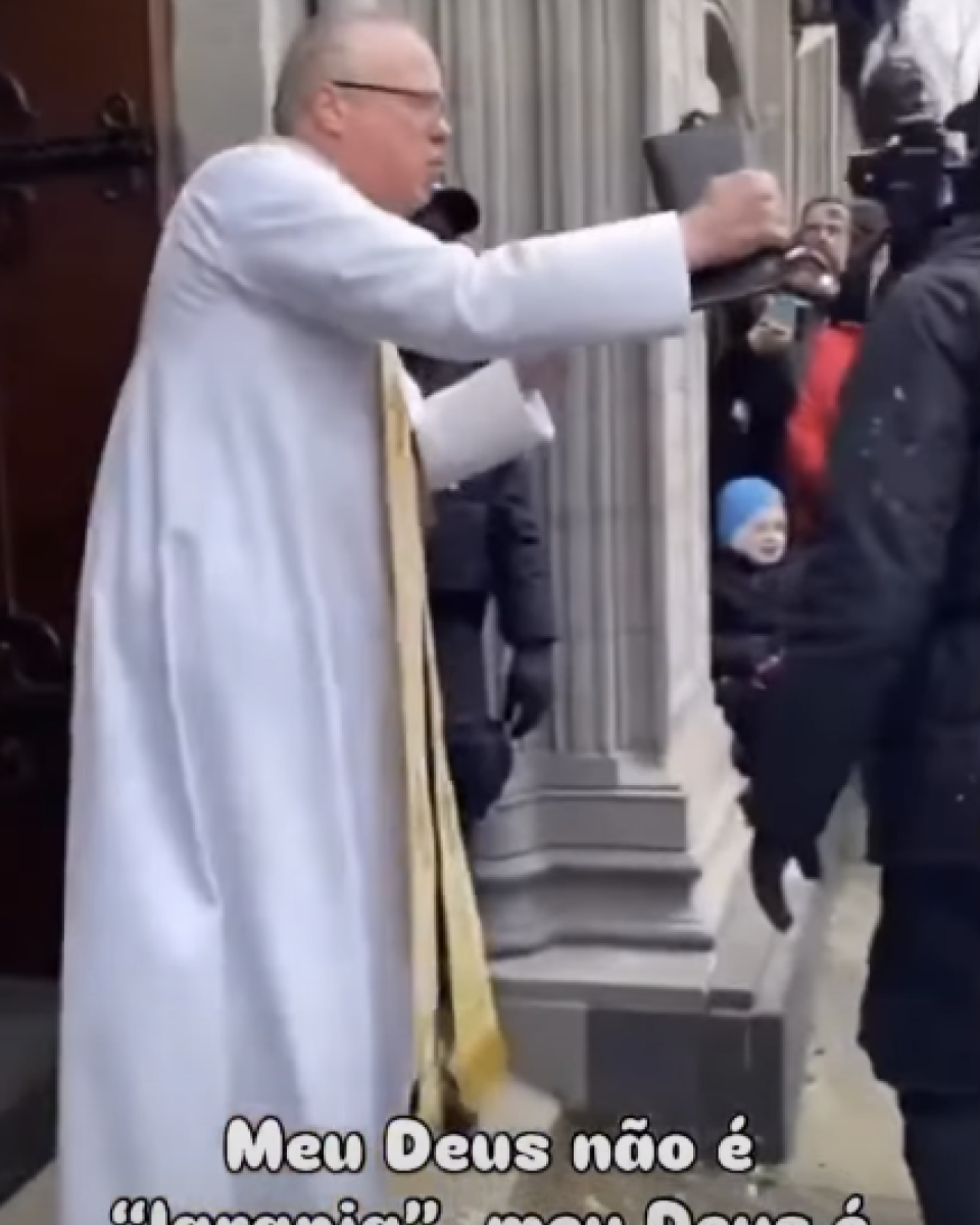 Imagem mostra uma cena externa em frente à entrada de um prédio, com homem idoso de pele clara, cabelo curto grisalho, usando óculos e veste longa branca com faixa vertical dourada, inclinado para a frente e estendendo o braço direito em direção a pessoas que estão à direita do enquadramento; ao redor, há outras pessoas parcialmente visíveis, incluindo indivíduos com roupas escuras. A fachada tem colunas e uma porta escura aberta, e na parte inferior da imagem aparece um texto sobreposto parcialmente visível que diz ‘Meu Deus não é laranja, meu Deus é amor’.