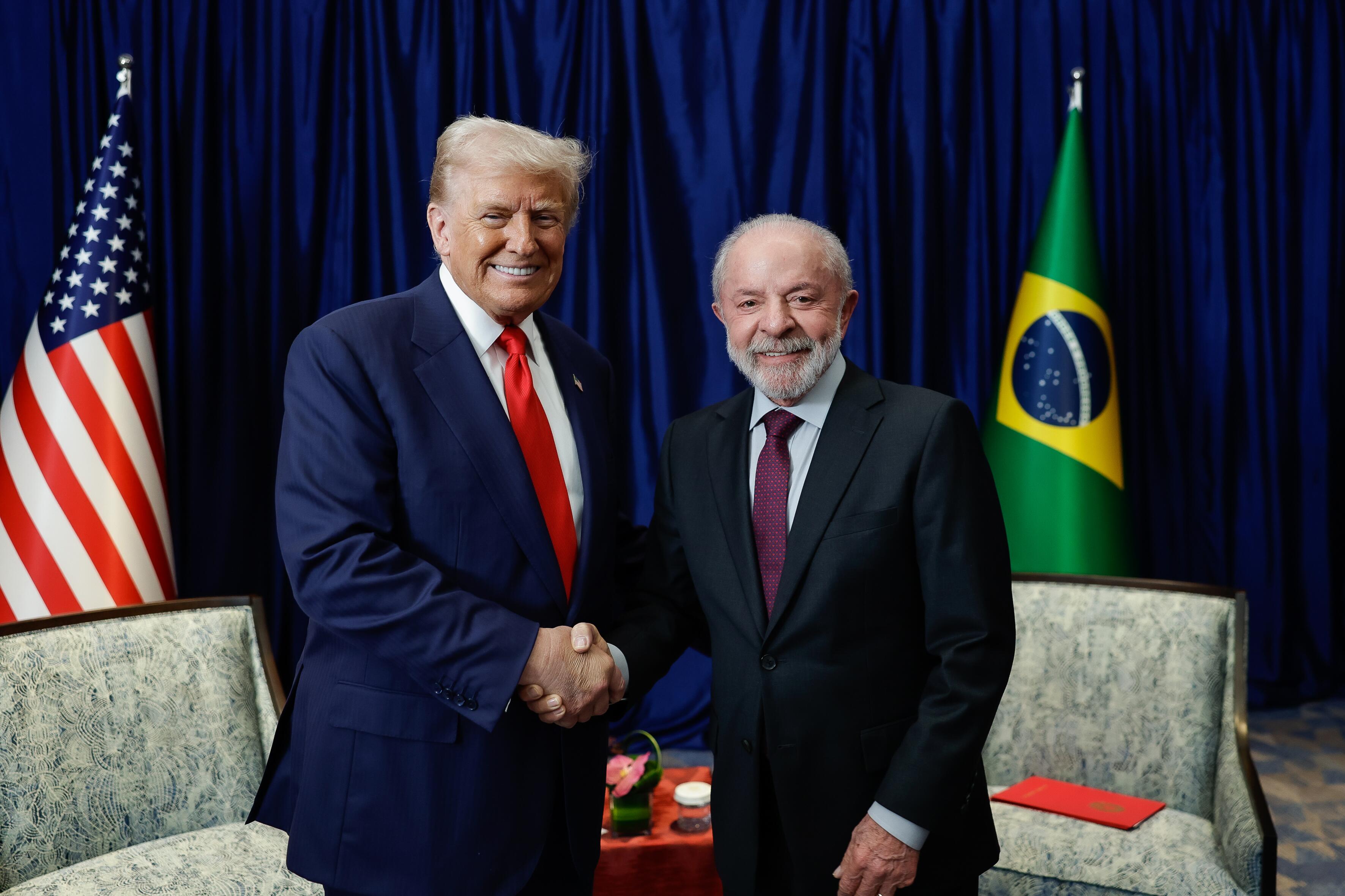 Imagem em formato horizontal mostrando os presidentes Donald Trump, dos Estados Unidos, e Lula, do Brasil, em pé, lado a lado, apertando as mãos no centro da cena. Ambos vestem ternos escuros; Trump está à esquerda e usa terno azul-escuro, camisa branca e gravata vermelha, tem cabelo claro penteado para trás e está sorrindo. Lula, à direita, usa terno preto, camisa branca e gravata em tom de vinho, tem cabelo curto grisalho, barba e bigode grisalhos, e também sorri. Ao fundo há uma cortina azul escura cobrindo toda a parede. À esquerda da imagem há uma bandeira dos Estados Unidos em um mastro. À direita há uma bandeira do Brasil em um mastro. Em primeiro plano, parcialmente visíveis, há duas cadeiras estofadas claras, uma de cada lado, e entre elas uma pequena mesa com um arranjo de flores e um objeto redondo. Sobre a cadeira à direita há um objeto retangular vermelho apoiado no assento.