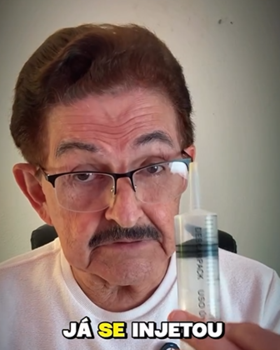 Imagem mostra um homem idoso, de pele clara, cabelo curto castanho escuro com áreas grisalhas, usando óculos de armação escura e camiseta clara, sentado diante de uma parede lisa de cor clara. Ele segura uma seringa transparente, posicionada próxima ao rosto, olhando diretamente para a câmera. Na parte inferior da imagem, há um texto em destaque que diz: ‘JÁ SE INJETOU’.