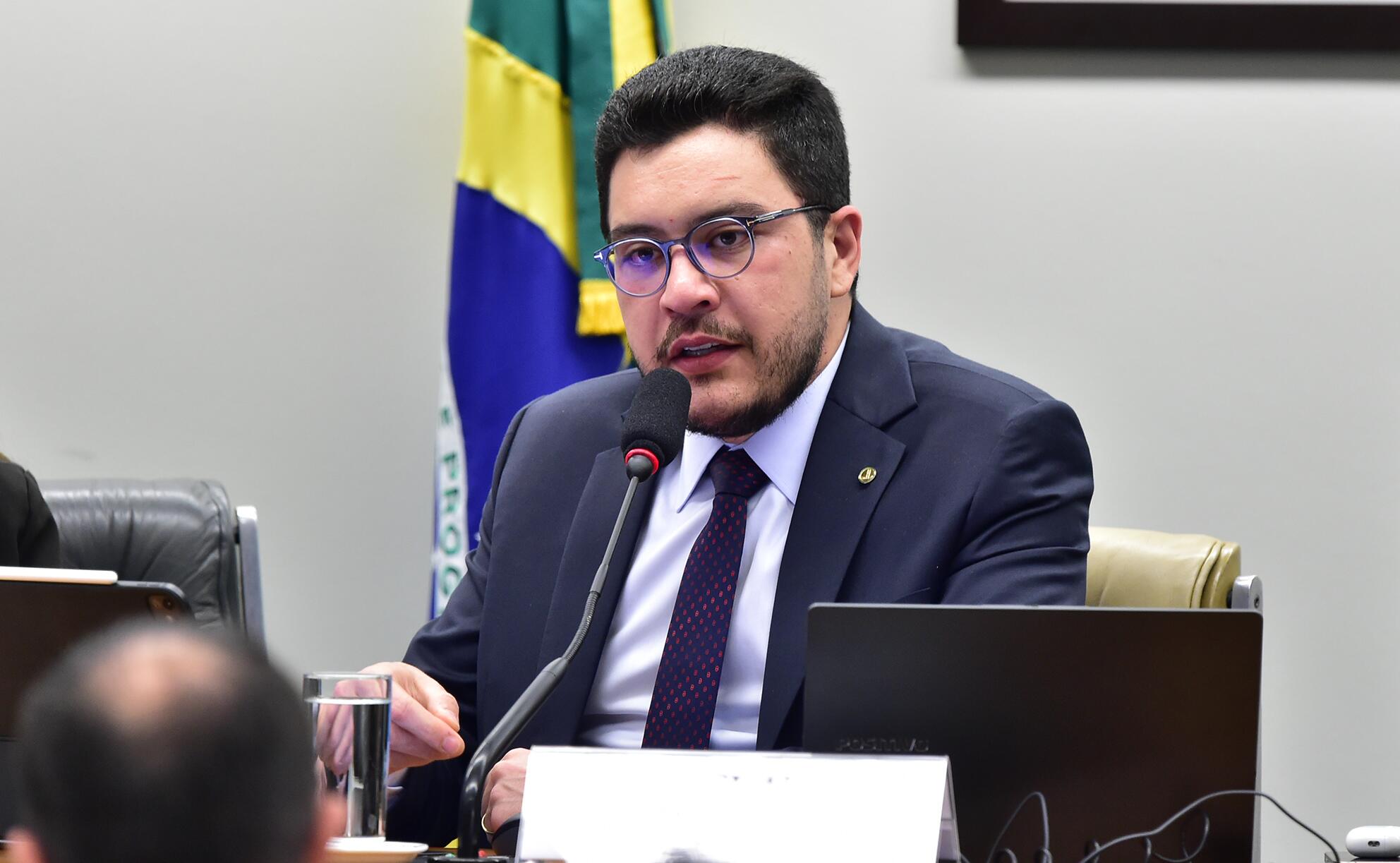 The image shows Congressman Jadyel Alencar — a man with light skin, short dark hair, and a short trimmed beard, wearing dark-framed glasses. He is sitting in front of a microphone and is dressed in a navy blue suit, a white shirt, and a dark tie with small dots. On the table in front of him, there is a glass of water, an open laptop, and a partially visible identification plate. In the background, a light-colored wall and part of a Brazilian flag in a vertical position can be seen, in a setting that suggests a meeting or an official session.