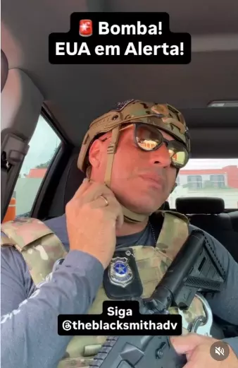 A imagem mostra um homem de pele clara sentado no banco do motorista de um carro, usando capacete tático camuflado, óculos escuros espelhados e colete tático camuflado sobre uma camiseta de manga longa cinza. Ele segura com a mão esquerda a alça do capacete próxima ao rosto e, com a mão direita, apoia um objeto escuro semelhante a um equipamento ou arma longa apoiado no corpo; no peito, há um distintivo circular preso ao colete. O interior do veículo é visível, com parte do volante e da porta, e ao fundo aparecem construções externas desfocadas. Na parte superior da imagem, há um texto gráfico que diz ‘Bomba! EUA em Alerta!’, e na parte inferior aparece o texto ‘Siga @theblacksmithadv’.