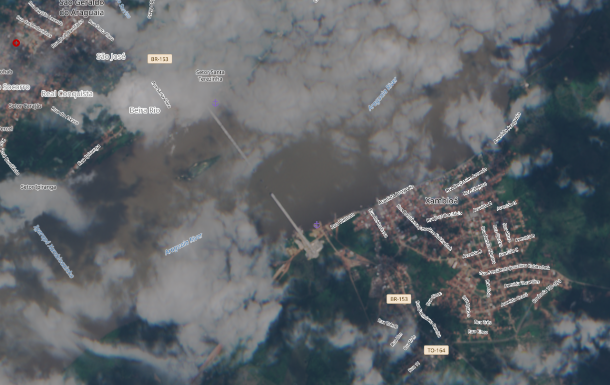 A imagem mostra uma visualização de satélite da região do rio Araguaia, com destaque para as cidades de Xambioá, no Tocantins, e São Geraldo do Araguaia, no Pará, separadas pelo curso do rio. É possível identificar áreas urbanas, vias sinalizadas com nomes de ruas e rodovias como a BR-153 e a TO-164, além de extensas áreas de vegetação. Duas estruturas lineares claras indicam a presença de uma ponte parcialmente construída ligando as margens do rio, parcialmente encoberta por nuvens, enquanto o leito do Araguaia aparece com coloração escura e irregular, sugerindo variação de profundidade ou sedimentos.