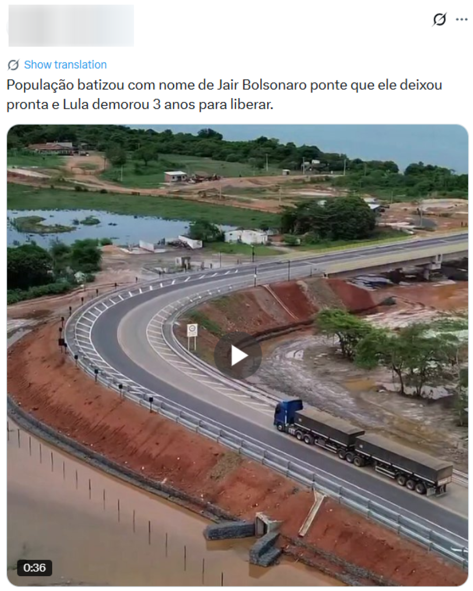 Post no X compartilha vídeo que mostra uma ponte rodoviária recém-construída sobre um curso d’água, com trechos de terra avermelhada exposta e pequenas construções ao redor. A via tem pista asfaltada com faixas de sinalização branca, guard-rails metálicos nas laterais e um acesso curvo que leva à travessia principal. Um caminhão de carga trafega sobre um dos acessos à ponte. Legenda do post diz: ‘População batizou com nome de Jair Bolsonaro ponte que ele deixou pronta e Lula demorou 3 anos para liberar’.