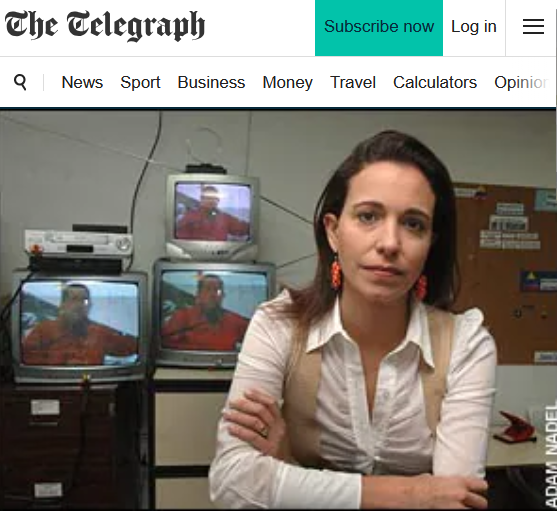 Imagem mostra María Corina Machado — mulher de pele clara e cabelo castanho escuro, preso para trás, sentada em uma sala com os braços cruzados, usando camisa branca de manga comprida, colete bege e brincos vermelhos. Ao fundo, há três televisores ligados exibindo a imagem de um homem vestindo roupa laranja, além de equipamentos eletrônicos e móveis de escritório