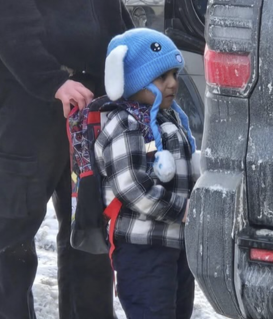 Imagem em formato vertical mostrando uma criança pequena em pé ao lado traseiro de um veículo. A criança está vestida com um casaco xadrez em tons de preto, branco e cinza, calça escura e usa uma mochila nas costas, com alças vermelhas e estampa colorida. Na cabeça, ela usa um gorro azul com abas laterais brancas, detalhes que lembram orelhas e dois cordões longos com pompons claros nas pontas. A criança está de perfil, voltada para a direita da imagem, olhando para a lateral do veículo. O carro é escuro e apresenta sujeira clara, semelhante a neve ou gelo, acumulada na lataria e no para-choque traseiro, com parte da lanterna vermelha visível. Atrás da criança, aparece parcialmente um adulto vestindo roupas escuras, com uma das mãos apoiada na parte superior da mochila. O chão é claro e irregular, também coberto por neve ou gelo. O ambiente sugere um local externo, frio, possivelmente um estacionamento ou área próxima a uma rua.