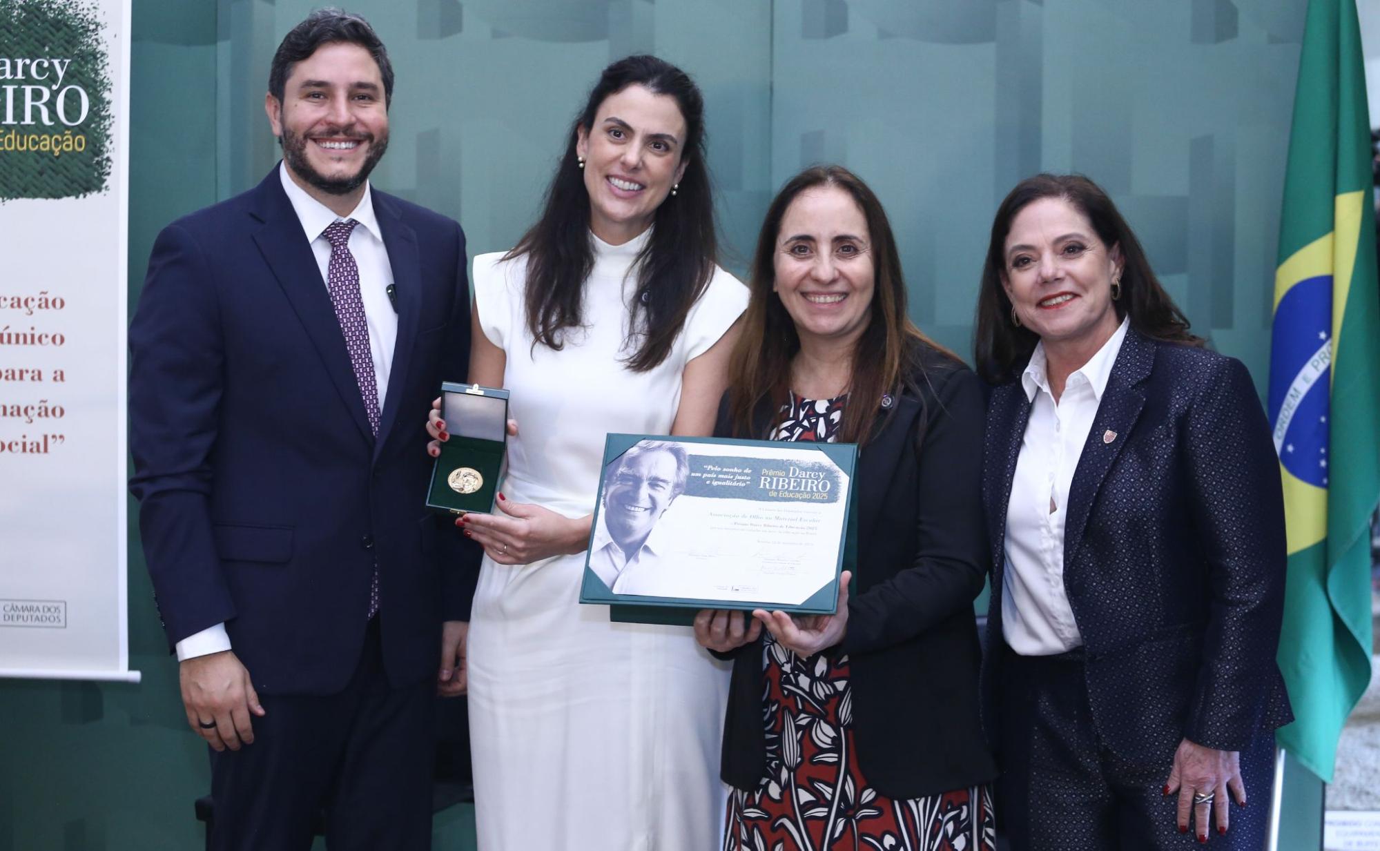 Grupo de quatro pessoas, um homem e três mulheres, posam para foto sorrindo em cerimônia oficial, em frente a um painel institucional e à bandeira do Brasil. No centro, Leticia Jacintho segura uma medalha ao lado da deputada Adriana Ventura, que exibe um certificado de premiação. Ambas são mulheres brancas de cabelos castanhos, lisos e compridos. Leticia veste vestido branco e Adriana, um vestido estampado vermelho sob blazer preto.
