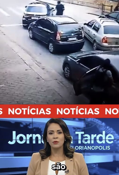 Imagem dividida em duas partes horizontais. Na parte superior, há uma imagem de câmera de segurança mostrando uma rua vista de cima e em ângulo inclinado. Vários carros estão parados ou alinhados próximos ao meio-fio, incluindo veículos nas cores preta, prata e branca. Um carro preto aparece mais próximo da câmera, com a porta aberta, e uma pessoa está parcialmente visível ao lado desse veículo. Mais à frente, outro carro está parado e uma pessoa aparece de pé próxima a ele, na pista. A calçada de pedras aparece à esquerda da imagem e a rua ocupa a maior parte do enquadramento. Na parte inferior da imagem, há um estúdio de telejornal. Uma apresentadora aparece do busto para cima, usando blazer bege sobre uma blusa clara, com cabelo castanho na altura dos ombros. Ao fundo, há um painel azul com o texto ‘Jornal da Tarde Florianópolis’. Acima da apresentadora, uma faixa vermelha atravessa a tela com a palavra ‘NOTÍCIAS’ repetida em letras brancas.