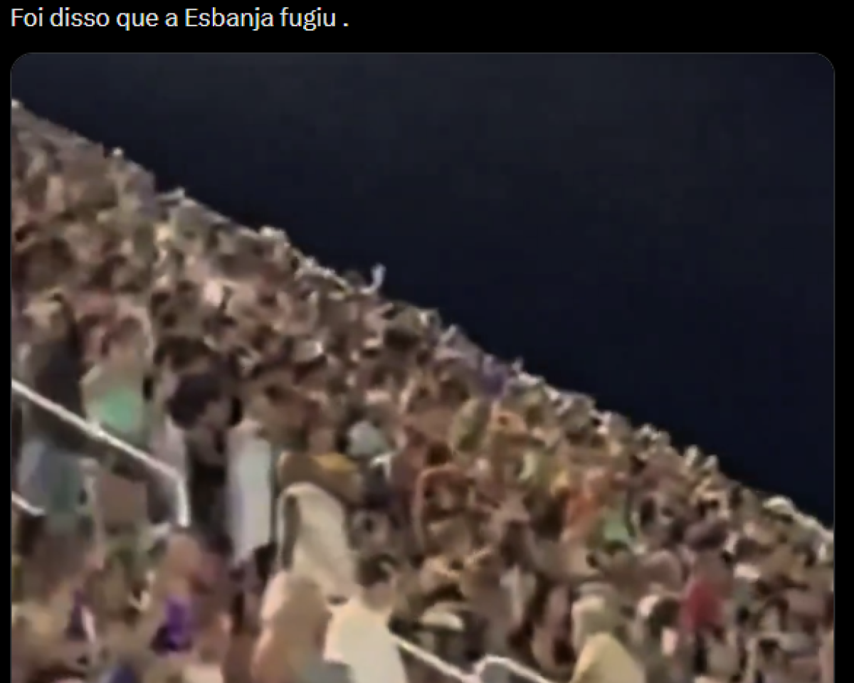 Imagem horizontal com fundo predominantemente escuro. Na parte superior, há uma faixa preta que diz ‘Foi disso que a Esbanja fugiu’. Abaixo do texto, ocupa quase toda a imagem uma grande arquibancada cheia de pessoas. A multidão aparece densa, com muitas mãos levantadas. Diversos cartazes e placas estão erguidos acima da cabeça das pessoas. As pessoas estão distribuídas em fileiras inclinadas, sugerindo um estádio ou arena.