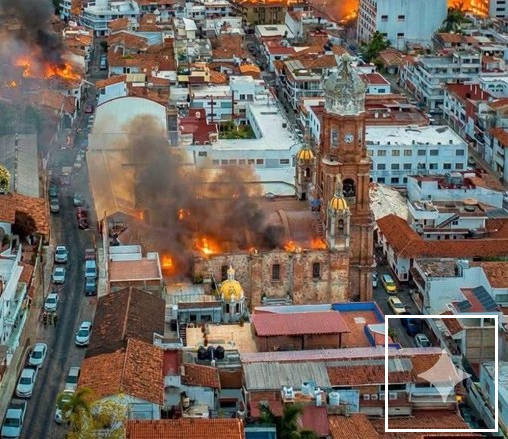Imagem aérea mostra conjunto de edificações urbanas, com destaque para igreja de torres e cúpulas claras no centro. O prédio aparece tomado por chamas e fumaça, que se espalha sobre os telhados ao redor. Ruas estreitas, casas de telhado cerâmico e prédios próximos cercam a área afetada pelo incêndio. Um quadrado com borda branca no canto inferior direito da imagem destaca uma marca d’água que indica uso da ferramenta Gemini, do Google.