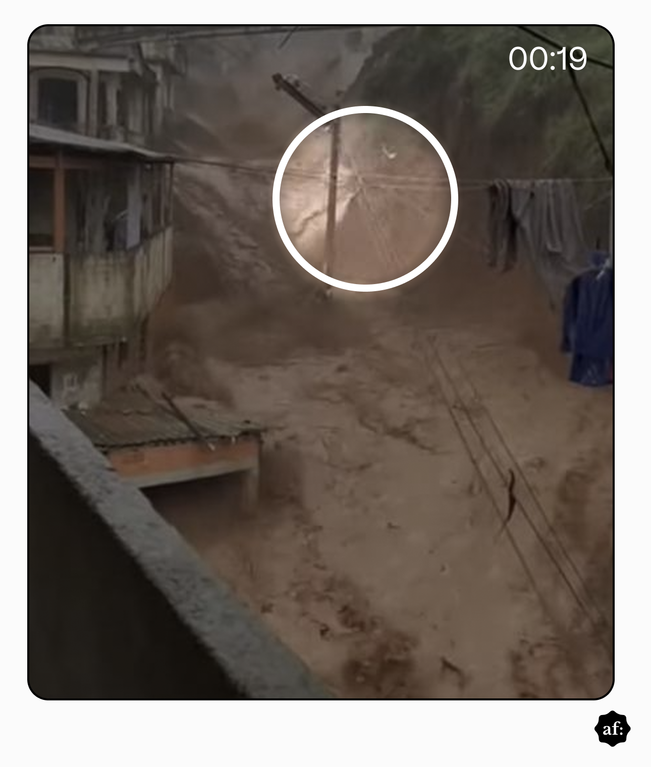 A imagem apresenta uma perspectiva em close de um deslizamento de terra ocorrendo em um corredor estreito entre construções. Uma torrente de lama e detritos marrons preenche o espaço central, descendo com força de uma área mais elevada ao fundo. À esquerda, vê-se a parede lateral e a varanda de uma casa de alvenaria cinza. À direita, um varal com peças de roupa escuras está pendurado, permanecendo imóvel enquanto a lama passa logo abaixo. Um círculo branco no centro da imagem destaca um poste de energia ou viga que parece estar inclinado ou sendo atingido pelo fluxo, e que não há um ‘final’ para o varal. Vários fios atravessam a cena de forma desordenada. No canto inferior esquerdo, parte de um telhado de telhas onduladas está parcialmente submerso pela terra. No canto superior direito, o cronômetro marca '00:19'.