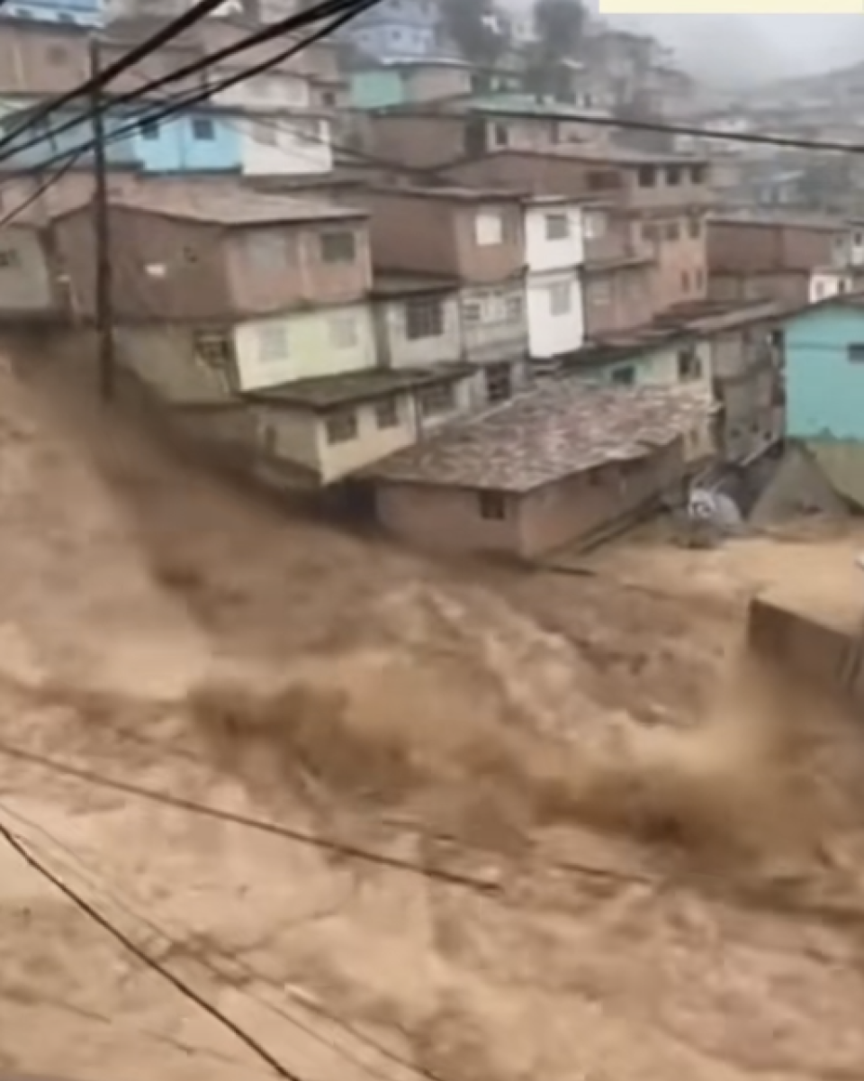 Imagem mostra área urbana em encosta. Várias casas simples e muito próximas umas das outras aparecem ao fundo. Na parte inferior da imagem, uma grande quantidade de água barrenta avança com força, carregando lama. Fios elétricos atravessam a cena em primeiro plano. O ambiente parece encoberto por chuva.