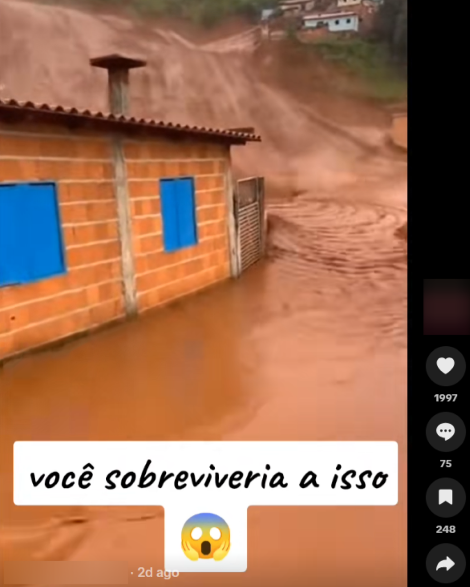 Casa de alvenaria com paredes de tijolo aparente e janelas azuis aparece parcialmente cercada por água barrenta em movimento. Ao fundo, encosta de terra úmida indica deslizamento, enquanto lama invade terreno ao redor da construção. Sobre o vídeo, há o texto ‘você sobreviveria a isso’, acompanhado de hashtags relacionadas a barragem, desastres e Minas Gerais.