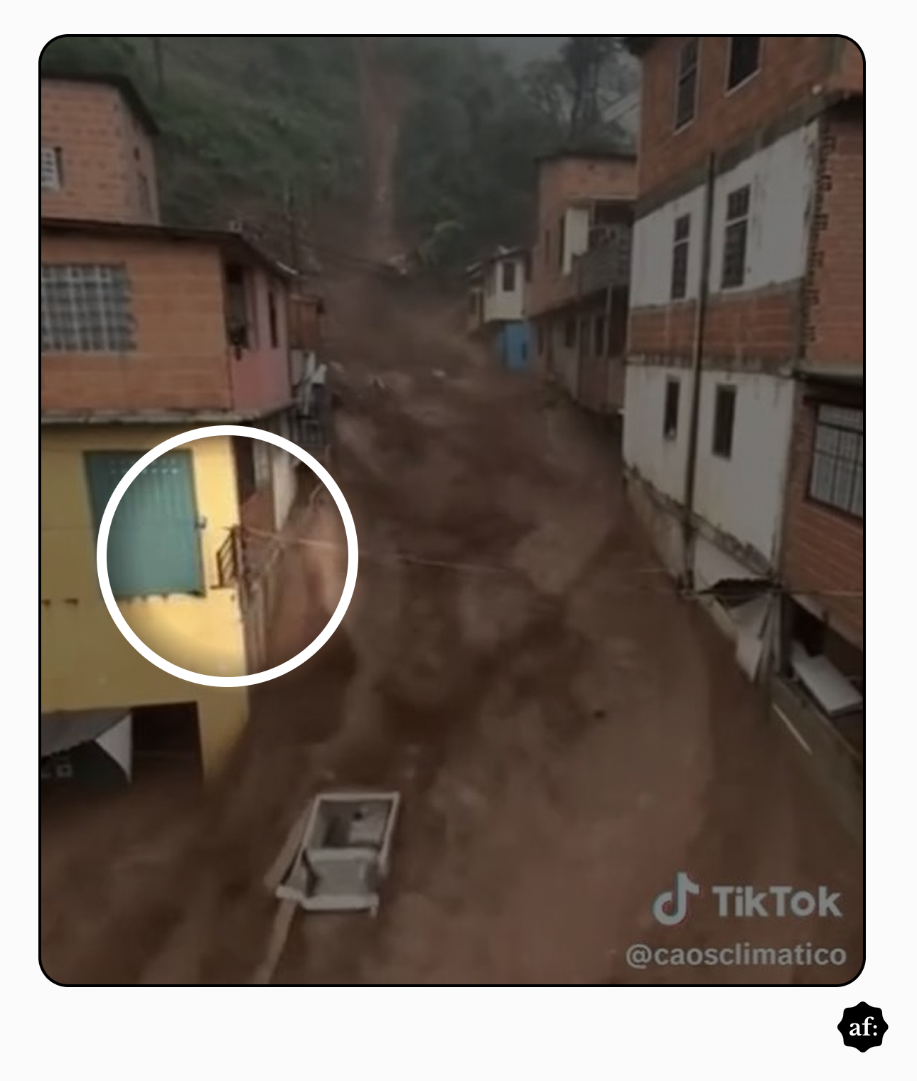 Rua estreita em área urbana construída em encosta, com casas de alvenaria em tons de bege, vermelho e branco distribuídas dos dois lados. A via está tomada por água barrenta em forte correnteza, que desce pelo declive entre as residências. Um círculo branco destaca a fachada de uma casa amarela com porta azul e um portão deslocado. No centro inferior da imagem, há um objeto retangular semelhante a uma caixa ou estrutura de concreto sendo arrastado pela água. No canto inferior direito, aparece a marca do TikTok e o nome de usuário ‘@caosclimatico’.