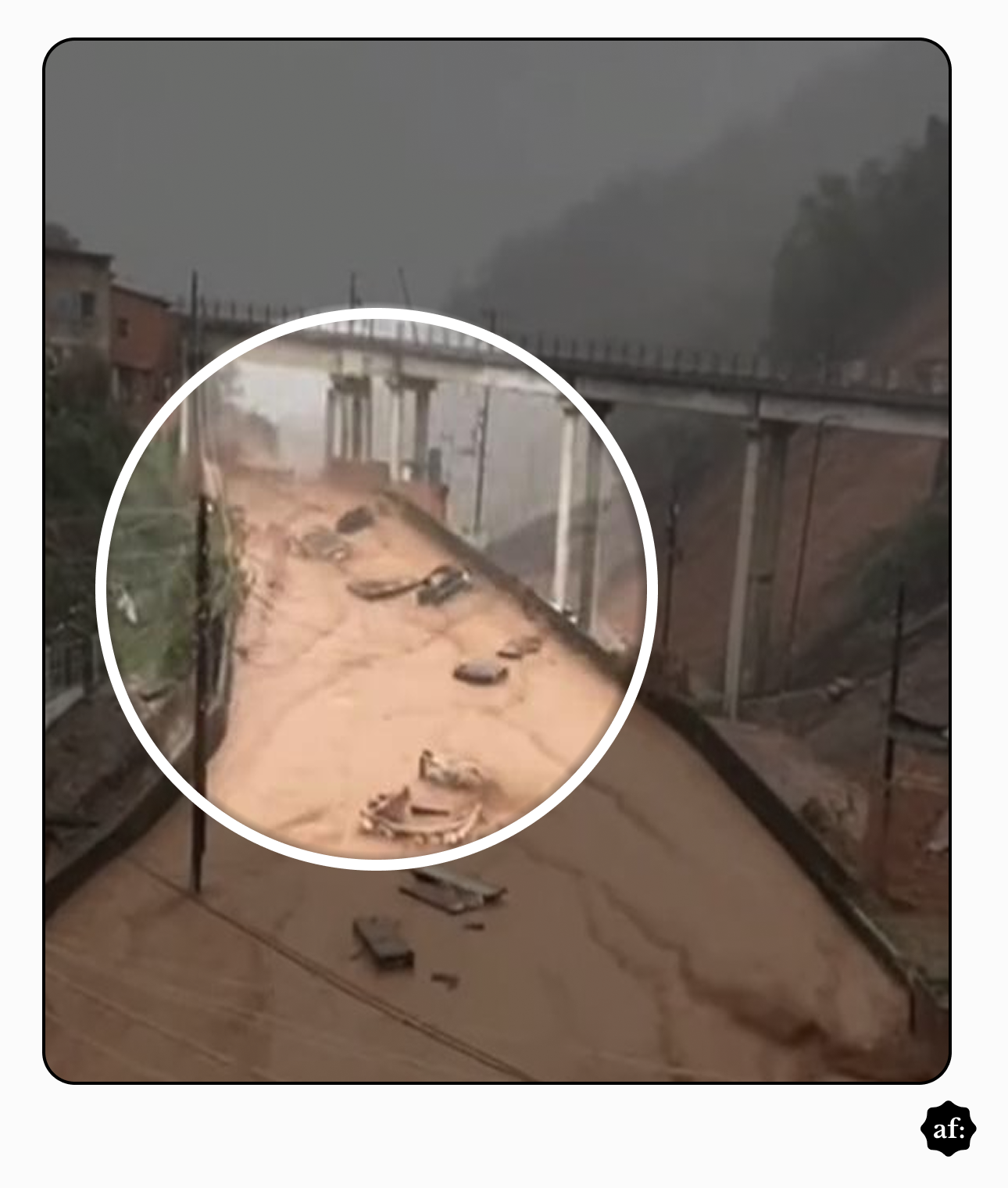 Via urbana alagada por água barrenta em forte correnteza, cercada por muros e encostas com vegetação. Ao fundo, há um viaduto sustentado por pilares de concreto que atravessa a área. Um círculo branco destaca a parte central da rua, onde aparecem veículos e destroços parcialmente submersos e espalhados pela água. O céu está encoberto por neblina ou chuva, e postes com fiação elétrica acompanham o trajeto da via.  