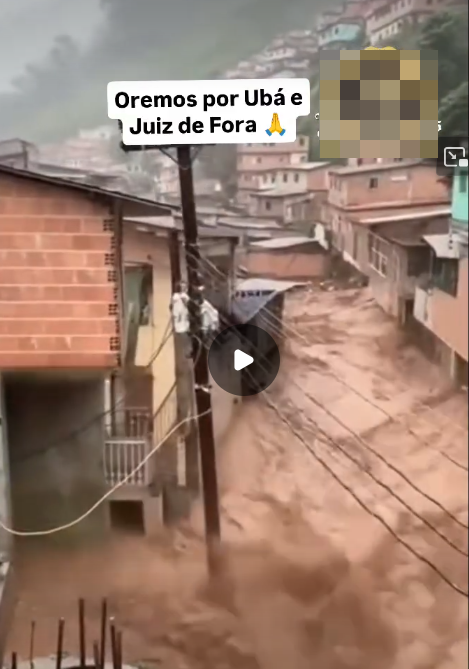 Cena de enchente em área urbana, com forte volume de água barrenta correndo por rua cercada por casas de alvenaria em tons de vermelho e bege, construídas em terreno inclinado. Um poste de energia atravessa o centro da imagem, e a água atinge a base das construções, formando correnteza intensa. Ao fundo, há moradias distribuídas pela encosta sob céu nublado. Sobre a imagem, aparece o texto ‘Oremos por Ubá e Juiz de Fora’, acompanhado de um emoji de mãos em oração.