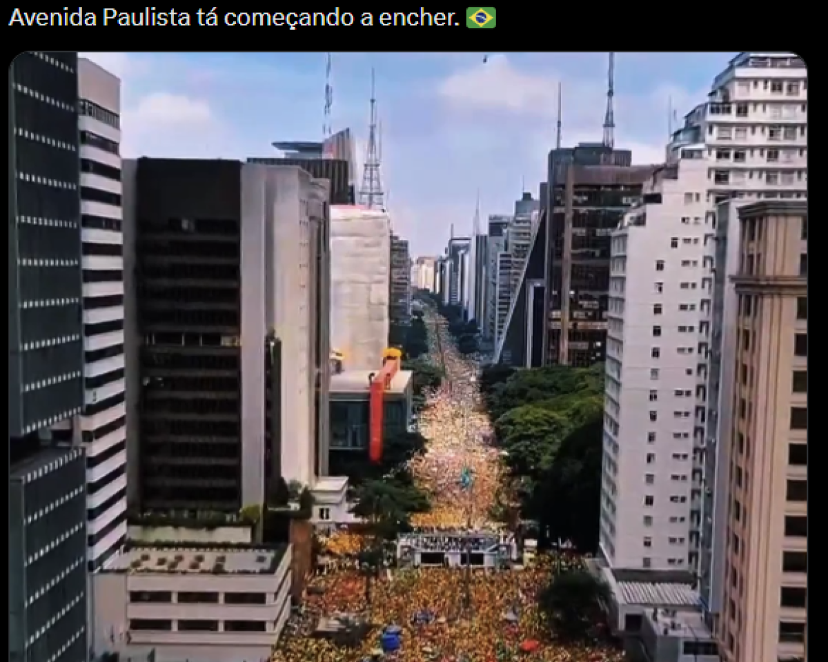 Publicação com texto: ‘Avenida Paulista tá começando a encher’, seguido de um emoji da bandeira do Brasil. Abaixo do texto, há uma fotografia aérea da avenida Paulista, em São Paulo. A via aparece tomada por uma grande multidão que ocupa toda a extensão da via visível na imagem. As pessoas formam uma faixa densa de cor predominantemente amarela ao longo do centro da avenida. De ambos os lados da rua há prédios altos, comerciais e residenciais, com fachadas em tons de branco, cinza e bege. Alguns edifícios possuem antenas e estruturas no topo. Árvores verdes acompanham a avenida em parte do trajeto. No ponto mais próximo da câmera, há uma estrutura montada sobre a via, semelhante a um palco ou pórtico. A multidão se estende até o horizonte, entre os prédios.