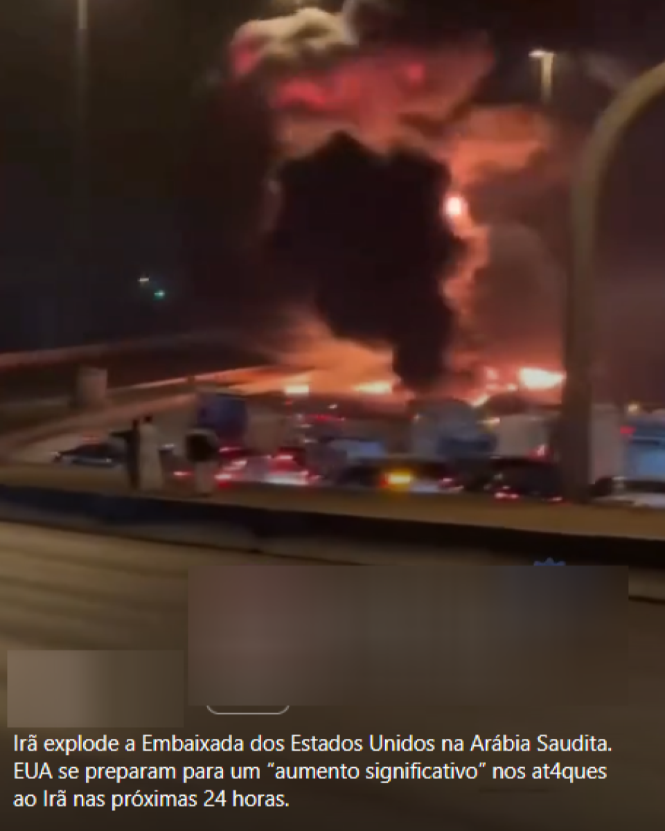 Imagem mostra área urbana à noite, com incêndio de grandes proporções ao fundo. Chamas alaranjadas e uma coluna densa de fumaça preta se elevam acima de construções e veículos estacionados, enquanto a cena é observada a partir de uma via elevada ou passarela. Na parte inferior, há um texto sobreposto que diz: ‘Irã explode a Embaixada dos Estados Unidos na Arábia Saudita. EUA se preparam para um “aumento significativo” nos ataques ao Irã nas próximas 24 horas.’