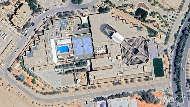 Imagem aérea capturada por satélite mostra a embaixada americana em Riade. O conjunto possui vários blocos interligados com telhados planos em tons claros, além de uma estrutura central com cobertura em formato piramidal com vigas aparentes. No interior do terreno, há uma piscina retangular azul, duas quadras esportivas lado a lado com marcações visíveis e áreas pavimentadas com palmeiras alinhadas. Ao redor do complexo, observam-se vias com carros estacionados, calçadas, árvores distribuídas ao longo das ruas e terrenos com solo arenoso.