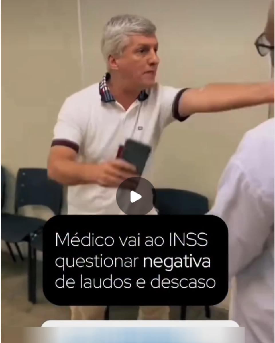 Homem de pele clara, cabelo curto grisalho e sem barba, vestindo camisa polo branca com detalhes escuros na gola e nas mangas, está em pé dentro de sala com cadeiras azuis alinhadas ao fundo. Ele segura um celular e estende o braço em direção a outro homem que aparece parcialmente de costas, usando camisa clara e óculos. Sobre o vídeo, há um texto que diz: ‘Médico vai ao INSS questionar negativa de laudos e descaso’.