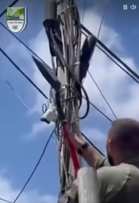 Imagem mostra homem de pele clara, cabeça raspada, visto de costas, usando farda clara, estendendo braço em direção a poste com vários cabos e equipamentos de telecomunicação presos à estrutura. Ele segura uma pá vermelha voltada para os fios. No topo do poste, há uma câmera de segurança, caixas e suportes metálicos ligados à fiação.