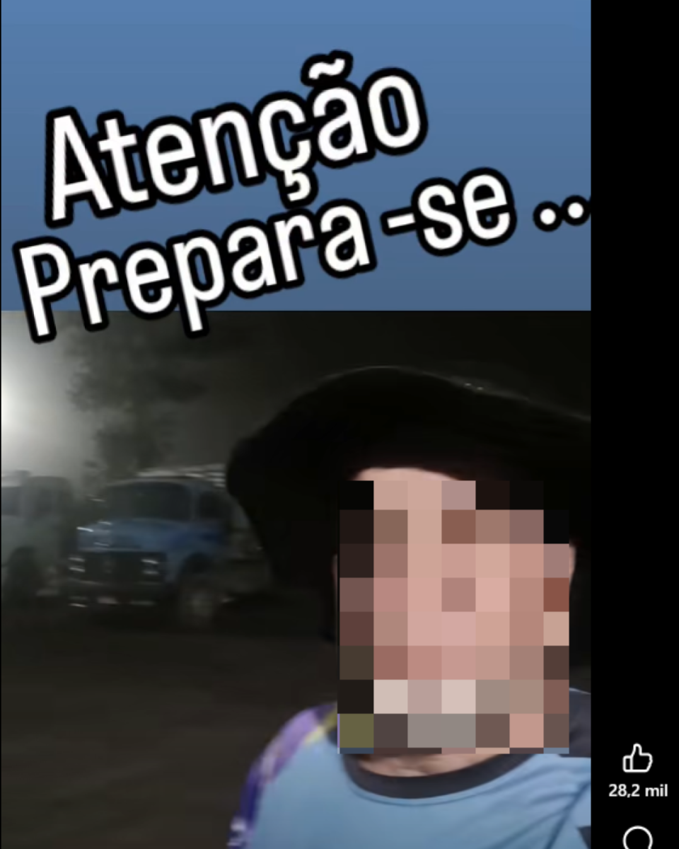 A imagem mostra um homem de pele clara, barba grisalha curta e usando um chapéu preto de aba larga, gravando um vídeo ao ar livre durante a noite. Ele veste uma camiseta azul com detalhes escuros nos ombros. Ao fundo aparecem caminhões estacionados e iluminação artificial que ilumina parcialmente o local, sugerindo um pátio ou área de parada de veículos pesados. Na parte superior da imagem há um texto em destaque que diz: ‘Atenção Preparara-se ..’.