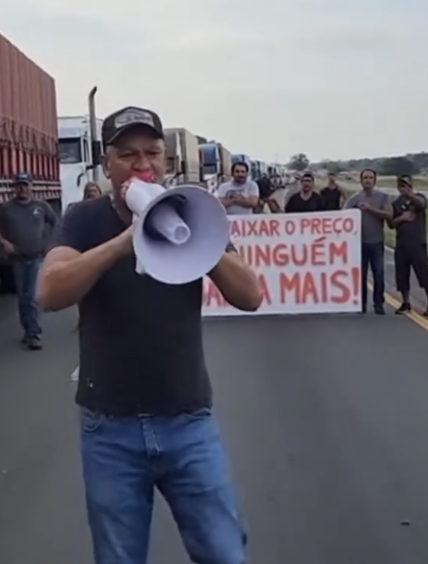 Print de uma publicação no TikTok que mostra um vídeo gerado por inteligência artificial retratando a cena de uma manifestação em uma rodovia. Em primeiro plano está um homem branco, usando um boné, vestindo uma camiseta preta e calça jeans segurando um megafone branco e vermelho na altura do rosto. Ao fundo aparecem outros homens segurando uma faixa branca com a seguinte frase em vermelho: ‘se não abaixar o preço, Lula, ninguém trabalha mais!’. Há ainda uma fileira de caminhões à esquerda da imagem.