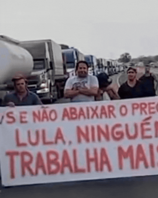 Trecho de um vídeo gerado por inteligência artificial mostra distorções nos rostos dos homens que aparecem na cena segurando uma faixa branca com a seguinte frase em vermelho: ‘se não abaixar o preço, Lula, ninguém trabalha mais!’. , além de um deles — que usa uma camisa xadrez — fazer movimentos incomuns com o corpo. Ao fundo há diversos caminhões parados, e o que está à esquerda da imagem tem um cano de descarga na porta do veículo.