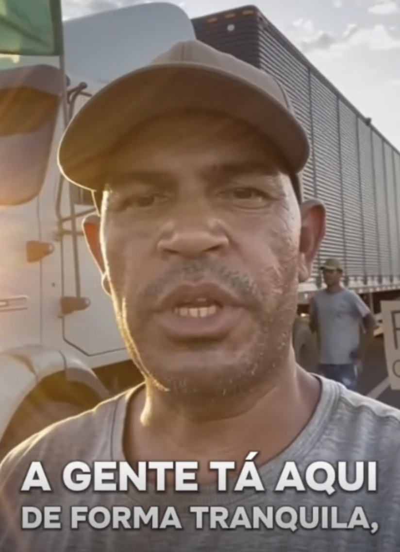 Print de uma publicação no TikTok que mostra um vídeo gerado por inteligência artificial retratando o momento em que um homem negro, usando um boné marrom e uma camiseta cinza relata uma suposta paralisação de caminhoneiros que estaria ocorrendo de ‘forma tranquila’. Ao fundo há um outro homem em pé e um caminhão parado.