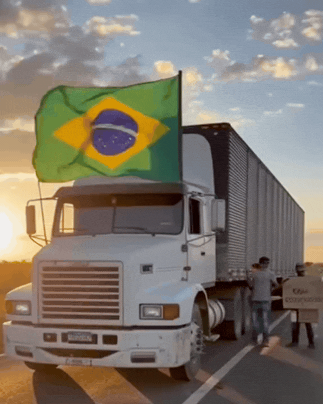 Trecho de um vídeo gerado por inteligência artificial mostra um caminhão parado em uma rodovia com uma bandeira do Brasil flamulando da direita para a esquerda. No céu azul, as nuvens brancas estão estáticas.