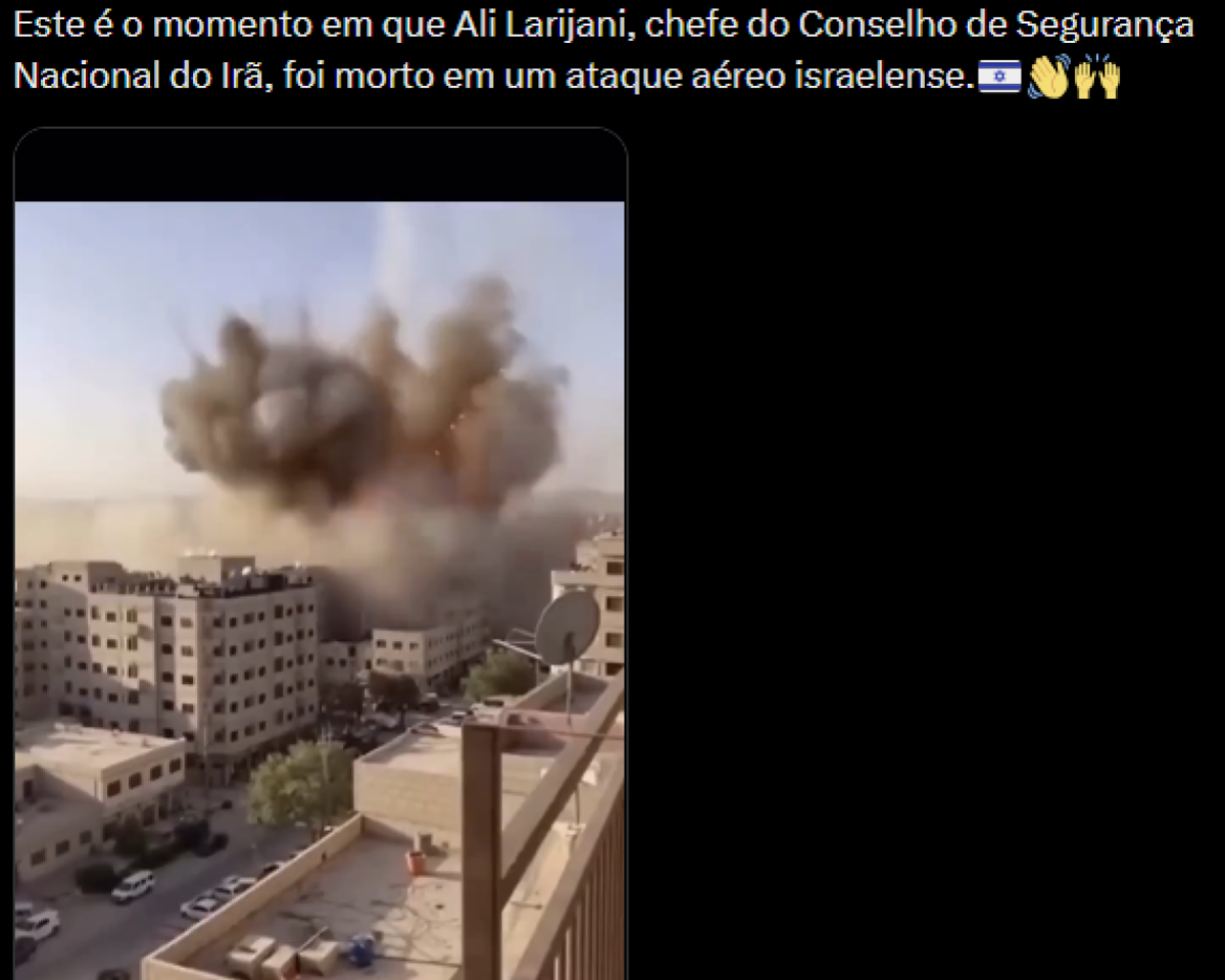 A imagem mostra uma publicação com fundo preto na parte superior, onde há um texto em branco que diz: Este é o momento em que Ali Larijani, chefe do Conselho de Segurança Nacional do Irã, foi morto em um ataque aéreo israelense, seguido de um emoji de bandeira de Israel e dois emojis de mãos levantadas. Abaixo do texto, há um vídeo em formato vertical. No quadro do vídeo, aparece uma área urbana com prédios de vários andares em tons claros, com janelas distribuídas pelas fachadas. No centro da imagem, há uma grande explosão formando uma nuvem densa de fumaça em tons de cinza e marrom que se expande acima dos edifícios. No primeiro plano, à direita, há uma sacada com corrimão e uma antena parabólica instalada. Na parte inferior, há uma rua com alguns carros estacionados. Ao fundo, a paisagem se estende com mais construções e terreno plano sob um céu claro.