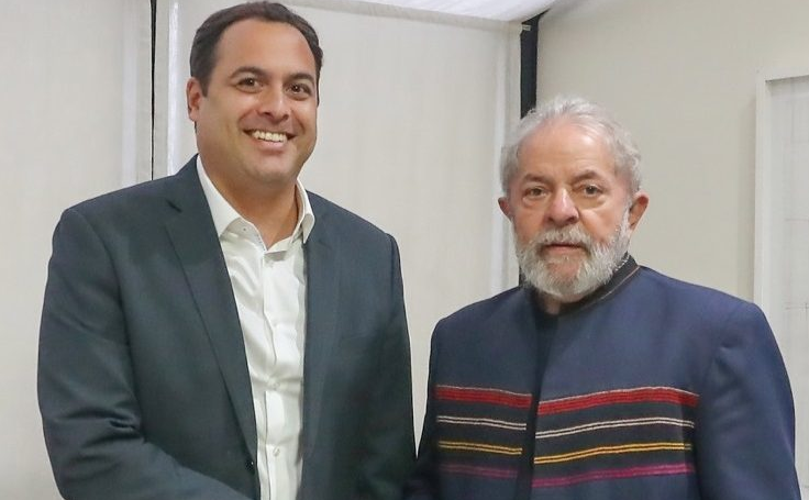 A imagem mostra dois homens posicionados lado a lado, de frente para a câmera, em um ambiente interno com paredes claras. À esquerda está Paulo Câmara, um homem de cabelo curto escuro, com um sorriso. Ele veste um terno escuro com uma camisa clara por baixo. Seu braço direito está estendido para frente, em um aperto de mão com Lula, que está à direita. O presidente tem cabelos e barba brancos. Ele veste uma peça de roupa escura com faixas horizontais em cores como vermelho e amarelo na altura do peito. Sua expressão é neutra. Ao fundo, há uma parede clara e uma janela com cortina branca parcialmente visível.