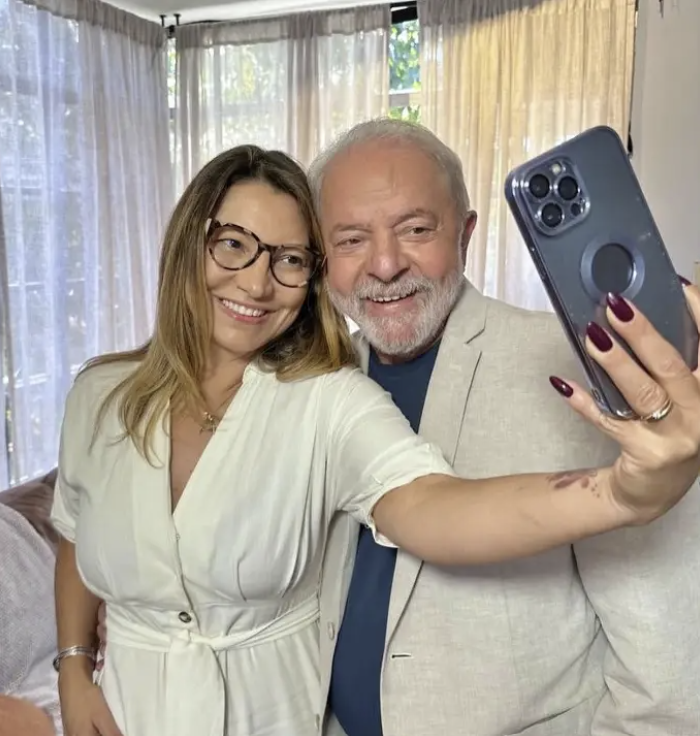 A imagem mostra Lula e Janja pessoas em pé, próximas uma da outra, sorrindo enquanto tiram uma selfie. Janja, à esquerda, tem cabelos longos e castanhos, usa óculos de armação escura e veste um vestido claro de manga curta com botões na parte frontal. Ela estende o braço direito para frente, segurando um celular com a câmera voltada para ambos. Lula, à direita, tem cabelos curtos e grisalhos, barba branca e veste uma camisa azul sob um blazer claro. Ele está levemente inclinado em direção à outra pessoa, com o rosto próximo ao dela. O celular aparece em primeiro plano, com três câmeras visíveis na parte traseira, e a mão que o segura tem unhas pintadas de cor escura. Ao fundo, há cortinas claras que cobrem janelas, permitindo a entrada de luz natural. À esquerda da imagem, há parte de um sofá ou assento claro.
