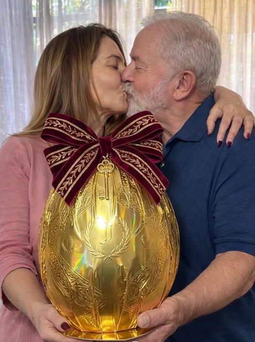 A imagem mostra Lula e Janja, com os rostos encostados, trocando um beijo. Ele, à direita, tem cabelos curtos e grisalhos, barba branca e veste uma camisa azul. Ela, à esquerda, tem cabelos longos e castanhos, veste uma blusa rosa e mantém um dos braços apoiado sobre o ombro da outra pessoa, com a mão visível nas costas. Ambos seguram, na altura do peito, um grande objeto em formato de ovo, de cor dourada, com detalhes ornamentais em relevo e um laço vermelho grande na parte superior. O objeto está apoiado em uma base e ocupa a parte central da imagem. Ao fundo, há cortinas claras que permitem a entrada de luz, com partes translúcidas e outras mais opacas. A iluminação é suave e uniforme.