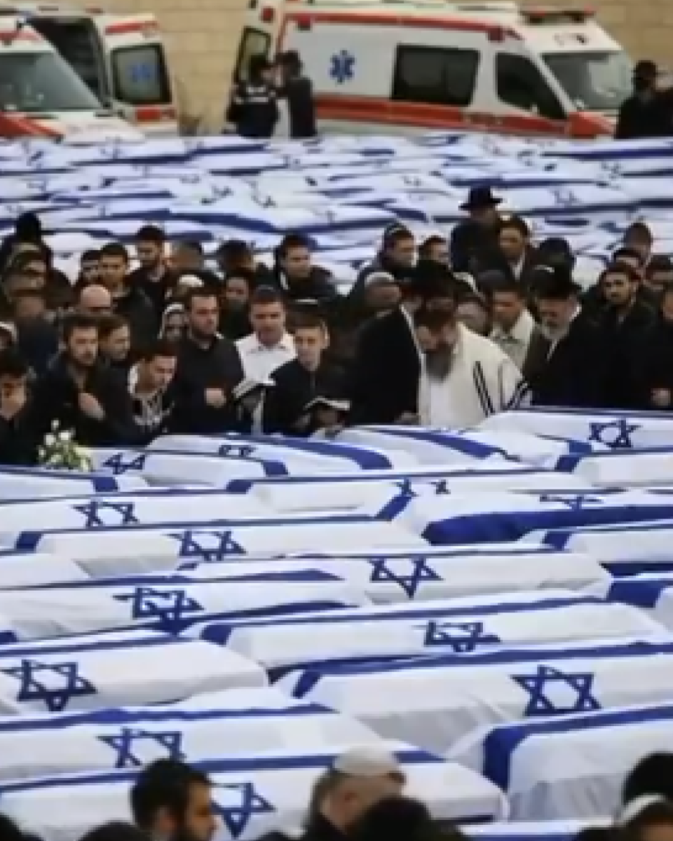 Imagem gerada por IA mostra uma grande quantidade de caixões dispostos lado a lado, todos cobertos por bandeiras de Israel, com a estrela de Davi visível. À frente dos caixões, há um grupo de pessoas reunidas, muitas vestidas de preto, algumas com a cabeça coberta, aparentando participar de uma cerimônia. No centro, um homem com vestimenta clara lê um texto, enquanto outros ao redor seguram livros ou observam em silêncio. Ao fundo, é possível ver ambulâncias estacionadas e mais pessoas agrupadas.