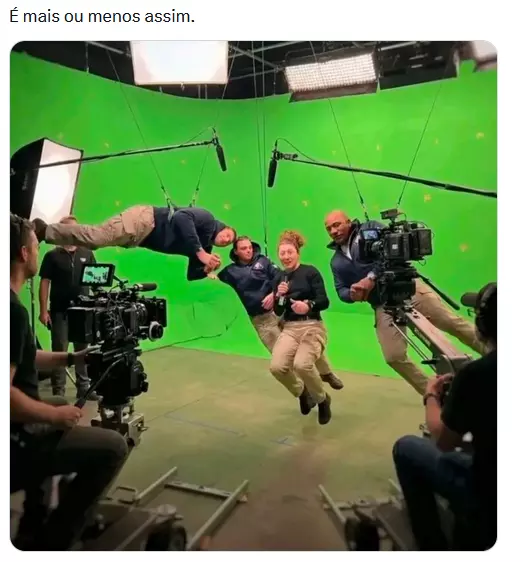 A imagem mostra um estúdio de gravação com fundo verde (‘chroma key’), onde quatro pessoas estão suspensas por cabos, simulando movimento no ar; eles vestem roupas em tons escuros e calças bege. Ao redor, há equipe de filmagem e equipamentos, incluindo câmeras profissionais, refletores e microfones suspensos por hastes. Um dos homens está na horizontal, enquanto os outros aparecem levemente inclinados, com expressões de esforço ou concentração. O cenário evidencia bastidores de produção audiovisual, com estruturas de iluminação e suporte visíveis.