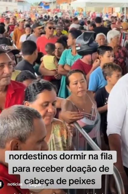 Grande aglomeração de pessoas em fila em ambiente externo, com homens e mulheres de diferentes idades e tons de pele vestindo roupas casuais em cores variadas. Em primeiro plano, uma mulher de pele clara, cabelo escuro preso e regata clara segura uma grade metálica, enquanto outras pessoas ao redor se aproximam. Entre a multidão, há um homem usando capacete de motocicleta e outros segurando crianças no colo. Ao fundo, a fila se estende por uma área ampla com construções simples. Na parte inferior da imagem, há um texto sobreposto que diz: ‘nordestinos dormir (sic) na fila para receber doação de cabeça de peixes’.