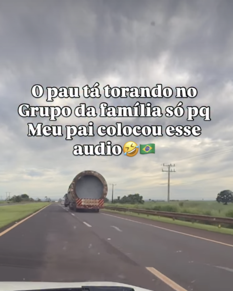 Estrada asfaltada vista de dentro de um veículo, com parte do painel aparecendo na parte inferior. À frente, há um caminhão transportando um objeto cilíndrico grande, semelhante a um tubo, ocupando a traseira do veículo. A estrada possui faixas de sinalização e segue em linha reta, com vegetação verde nas laterais e alguns postes de energia ao longo do caminho. O céu está nublado, com nuvens densas ocupando a maior parte da imagem. Ao fundo, há áreas abertas de campo e poucas árvores espalhadas. No centro da imagem, há um texto em letras grandes que diz: ‘O pau tá torando no Grupo da família só pq Meu pai colocou esse audio’, seguindo dos emojis de gargalhada e da bandeira do Brasil.